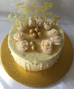 This is a Golden Wedding Anniversary Cake. It is a three layer, vanilla sponge. It is filled with vanilla buttercream and salted caramel. It is iced with vanilla buttercreamand has a white chocolate drip coat. It is decorated with cream sugar paste roses, gold-dusted chocolate balls and vanilla macarons.