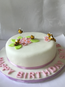 A pinapple flavoured cake covered with white icing and decorated with flowers and bees in sugar paste.